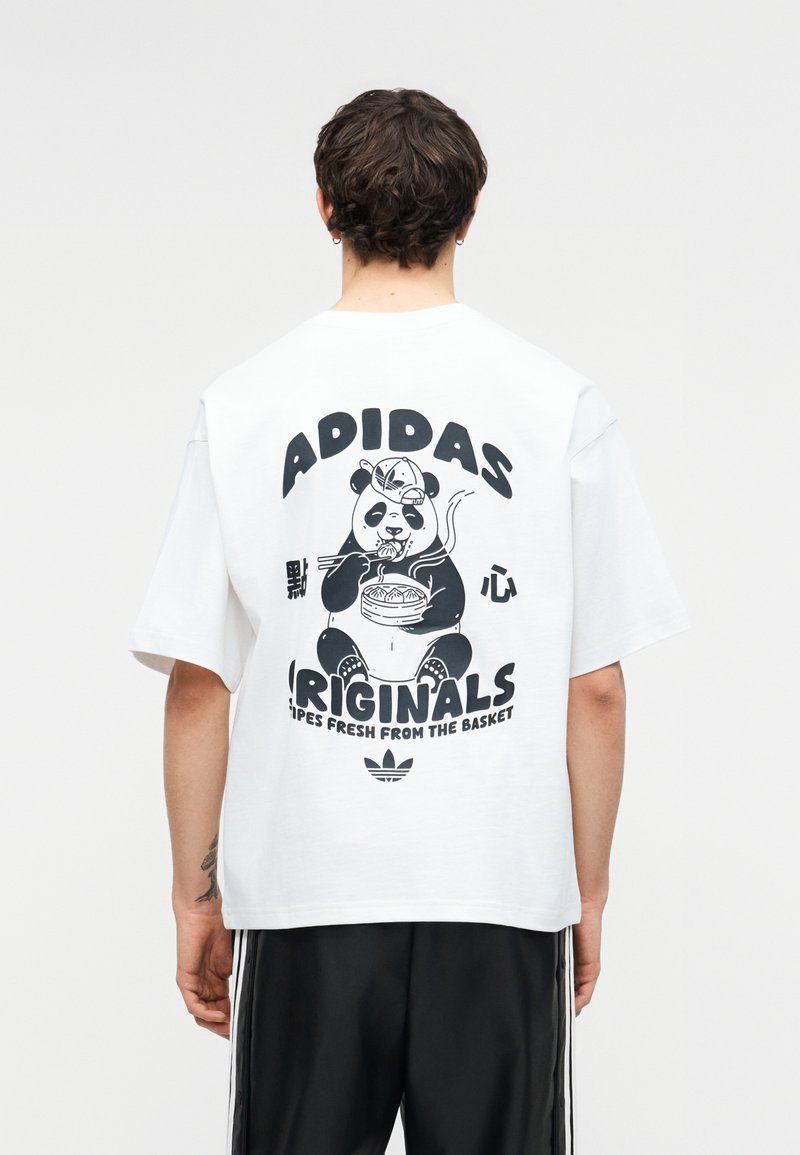 Osoba nosząca białą koszulkę Adidas z czarną grafiką pandy, jedząca makaron, z napisem „Adidas Originals” na plecach.