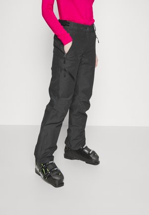 Person trägt schwarze Schneehose mit Reißverschlusstaschen und schwarze Skistiefel, steht mit einer Hand in der Tasche vor einfarbigem Hintergrund.