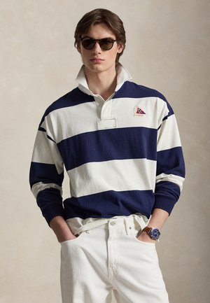 Młody mężczyzna w granatowo-białej pasiastym koszulce rugby, białych spodniach, brązowym skórzanym zegarku na rękę i ciemnych okularach przeciwsłonecznych, stoi z rękami w kieszeniach.