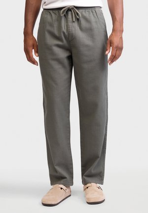 Mężczyzna w oliwkowozielonych spodniach z trokami i beżowych wsuwanych butach, stojący z ramionami swobodnie opuszczonymi wzdłuż ciała na jednolitym tle.