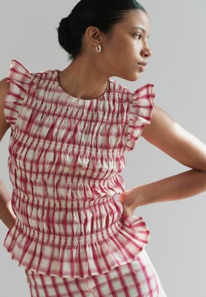 Žena s tmavými vlasmi vo vrkoči nosí červené a biele volánové kárované tielko bez rukávov a zodpovedajúcu sukňu, pózuje s rukami na bokoch, hľadí doprava.