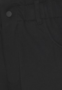 Närbild av svart tyg med synliga sömmar, en rund svart knapp och en del av en ficksöm på ett plagg.