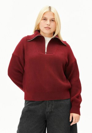 Jeune femme aux longs cheveux blonds portant un pull en maille rouge avec un col à fermeture éclair partielle et un pantalon noir à jambes larges, debout les mains dans le dos.