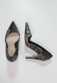 Svarta och grå sko med ormskinnsmönster, högklackade med spetsig tå och öppna sidor. Klacken är smal och cirka 10 cm hög.