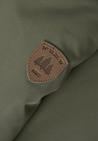 Fondo di tessuto verde oliva con una toppa in pelle marrone che presenta tre pini e la scritta "N° 18.25. W NMT".