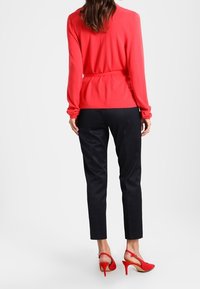 Top rojo de manga larga con cinturón, combinado con pantalones negros texturizados y zapatos de tacón rojos. Vista trasera, mostrando los detalles y el ajuste de la prenda.