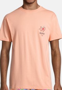 Camiseta de algodón melocotón con un gráfico en el pecho de un pulpo rojo y una marca de texto en gris oscuro. Manga corta y corte relajado.