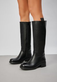 Bottes noires en cuir jusqu'aux genoux avec une texture lisse, un bout angle et un talon carré. Elles sont dotées d'une fermeture éclair et d'une languette à l'arrière.