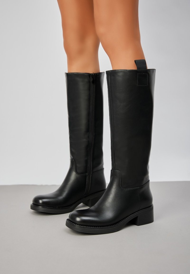 Bottes noires en cuir jusqu'aux genoux avec une texture lisse, un bout angle et un talon carré. Elles sont dotées d'une fermeture éclair et d'une languette à l'arrière.