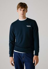 Marineblaues Sweatshirt mit rundem Hals, strukturiertem Stoff und einem weißen "RED BULL RACING"-Logo auf der linken Brust. Über einem weißen Hemd getragen.
