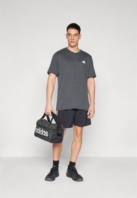 Szürke pamut póló környakkal és fehér logóval, fekete rövidnadrággal párosítva. Egy fekete sporttáskát tart, melyen fehér "adidas" felirat található.