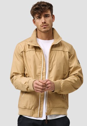 Ung mann med krøllete hår som har på seg en beige zip-up jakke over en hvit skjorte, står med hendene foldet mot en enkel grå bakgrunn.