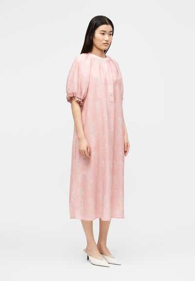 Γυναίκα που στέκεται, φορώντας ένα φαρδύ, ανοιχτορόζ φόρεμα μέχρι το γόνατο με φουσκωτά μανίκια και λευκές μυτερές χαμηλές γόβες σε μονόχρωμο φόντο.