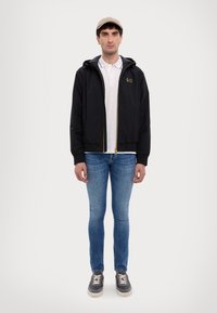 Black jacket with hood, zippered front; gold accents. White polo underneath; blue jeans and gray sneakers complete the outfit.