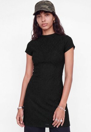 Le mannequin porte une robe noire ajustée à manches courtes, une casquette camouflage, des créoles, des bracelets et des bagues, sur un fond de studio blanc.
