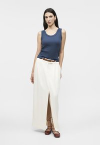 Femme portant un haut bleu sans manches, une jupe blanche taille haute avec une fente à l'avant, une ceinture marron et des chaussures plates marron à lacets, debout devant un fond uni.