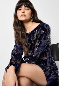 Granatowa sukienka z cekinami, z długimi rękawami, dopasowanym kształtem i lekko odkrytymi ramionami. Gładka tekstura z metalicznymi akcentami.