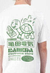 Witte t-shirt met een groene afbeelding van een schildpad en een elektronisch apparaat, met tekst in het Japans en het Engels. Het bedrukt ontwerp bevat details van circuits.