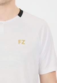 Valkoinen urheilu paita, jossa on musta kaulus, lyhyet hihat ja kultainen "FZ" -logo vasemmalla puolella. Pehmeä kangas, jossa on hienovarainen rakenne.