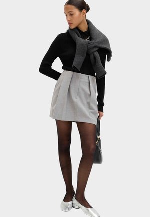 Sort ribbet sweater, grå plisseret mininederdel, gennemsigtige strømpebukser, sølvfarvede flade sko og et mørkegråt tørklæde draperet omkring skuldrene. Sort håndtaske.