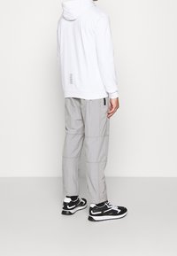 Bílá mikina s kapucí s potiskem na zádech, šedé lehké kalhoty a černé tenisky. Výrazné detaily zahrnují černé zipy na kalhotách a elegantní design obuvi.