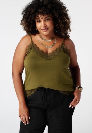 Vrouw met krullend haar die een olijfgroene top met kanten randje draagt en zwarte gestructureerde broek met gelaagde gouden kettingen en armbanden.