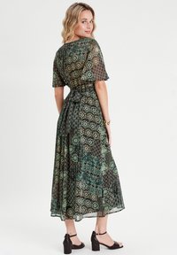 Robe maxi en tissu léger avec un motif floral et géométrique en vert, noir et beige. Taille nouée et manches courtes à volants.