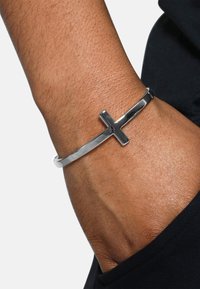 Metallarmbånd med en polert sølvfinish, som har et korsdesign. Båret på håndleddet, som fremhever en glatt, avrundet form.