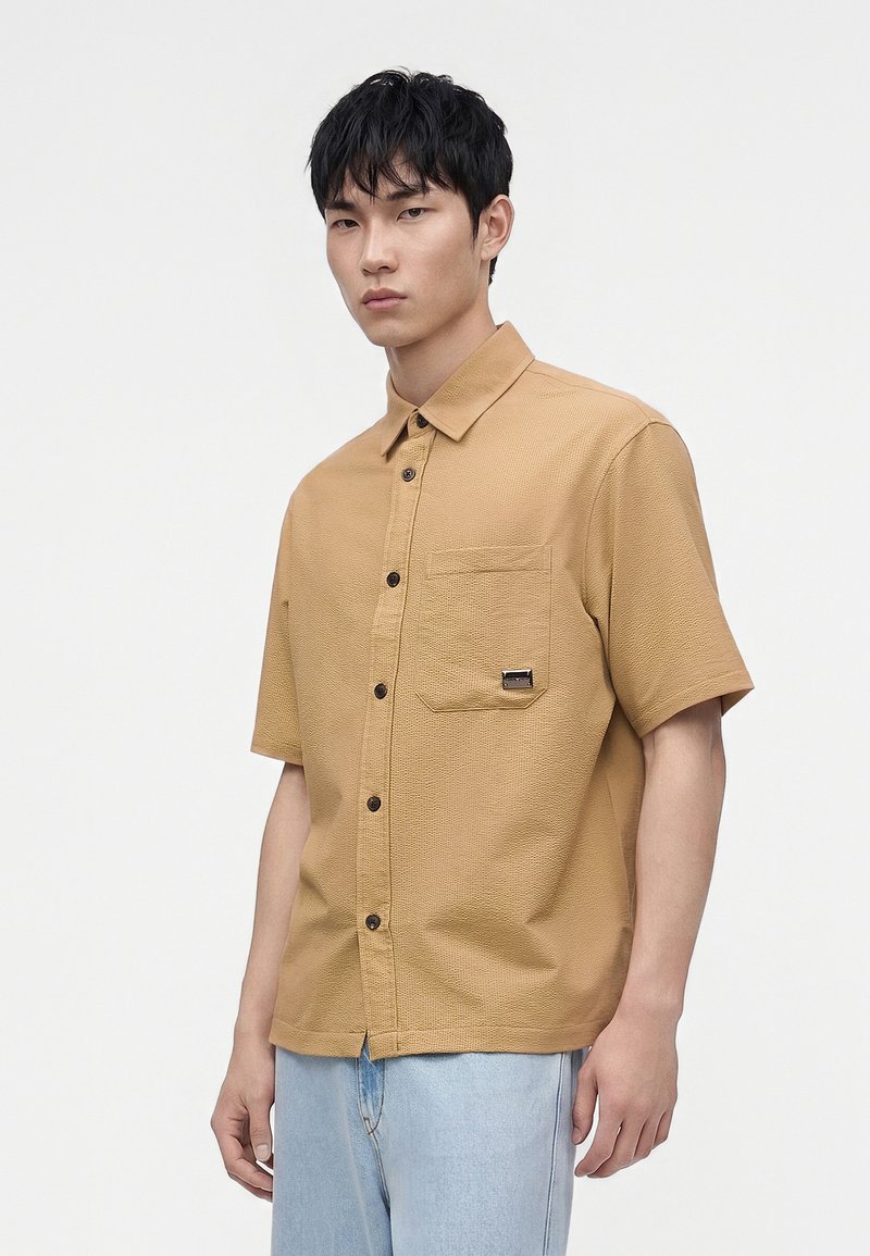 Jeune homme aux cheveux courts noirs porte une chemise beige à manches courtes boutonnée et un jean bleu clair sur un fond blanc uni.