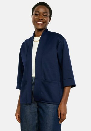 Lächelnde Frau, die eine marineblaue, offene Jacke mit Dreiviertelärmeln, ein weißes Hemd, eine goldene Halskette und blaue Jeans trägt.