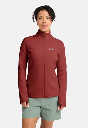 Frau trägt eine dunkelrote Jack Wolfskin Jacke mit Reißverschluss und hellgrüne Shorts, steht vor einem schlichten weißen Hintergrund.
