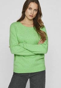 Zeleni pulover iz mehke, teksturirane tkanine z dolgimi rokavi ter rebrastimi manšetami in robom. V kombinaciji z temno sivimi hlačami.