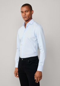 Muškarac u svijetloplavoj košulji na gumbe i crnim hlačama, stoji s neutralnim izrazom lica nasuprot jednostavne svijetle pozadine.