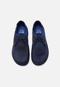 Ζεύγος μοκασίνια από μπλε σουέτ, με ταίριασμα κορδόνια και επενδεδυμένες μπλε εσωτερικές σόλες, εμφανίζονται από πάνω σε λευκό φόντο.