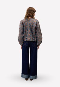 Frau mit welligem Haar, die ein langärmliges Oberteil mit floralem Muster und weiten, hellblauen, hochgekrempelten Jeans trägt, steht mit dem Rücken zur Kamera.