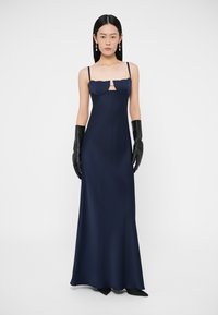 Tumši zila, līdz grīdai sniedzoša kleita ar piegulošu siluetu, dāļveida kakla izgriezumu un dekoratīvām atloku detaļām. Melni cimdi papildina tēlu.