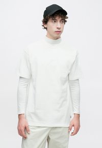 Άσπρη κοντομάνικη μπλούζα που φοριέται πάνω από μια λευκή μακρυμάνικη μπλούζα με διακριτικό λογότυπο, σε συνδυασμό με μαύρο καπέλο και ανοιχτόχρωμο παντελόνι.