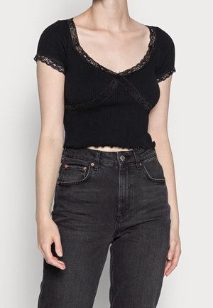 Frau trägt ein schwarzes Kurzarmoberteil mit Spitzenbesatz und hoch taillierte schwarze Jeans, steht vor einem schlichten hellen Hintergrund.