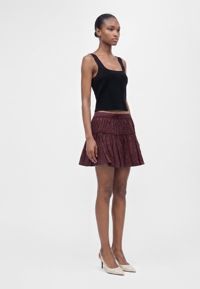 Femme debout portant un haut noir sans manches, une jupe bordeaux à œillets et des chaussures à talons beiges sur un fond clair uni.