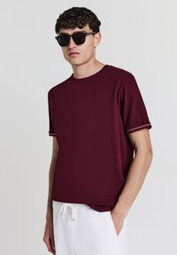 Giovane uomo con occhiali da sole scuri, una camicia a maniche corte bordeaux con cuciture bianche sulle maniche e pantaloni bianchi con cordoncino, con una mano in tasca.