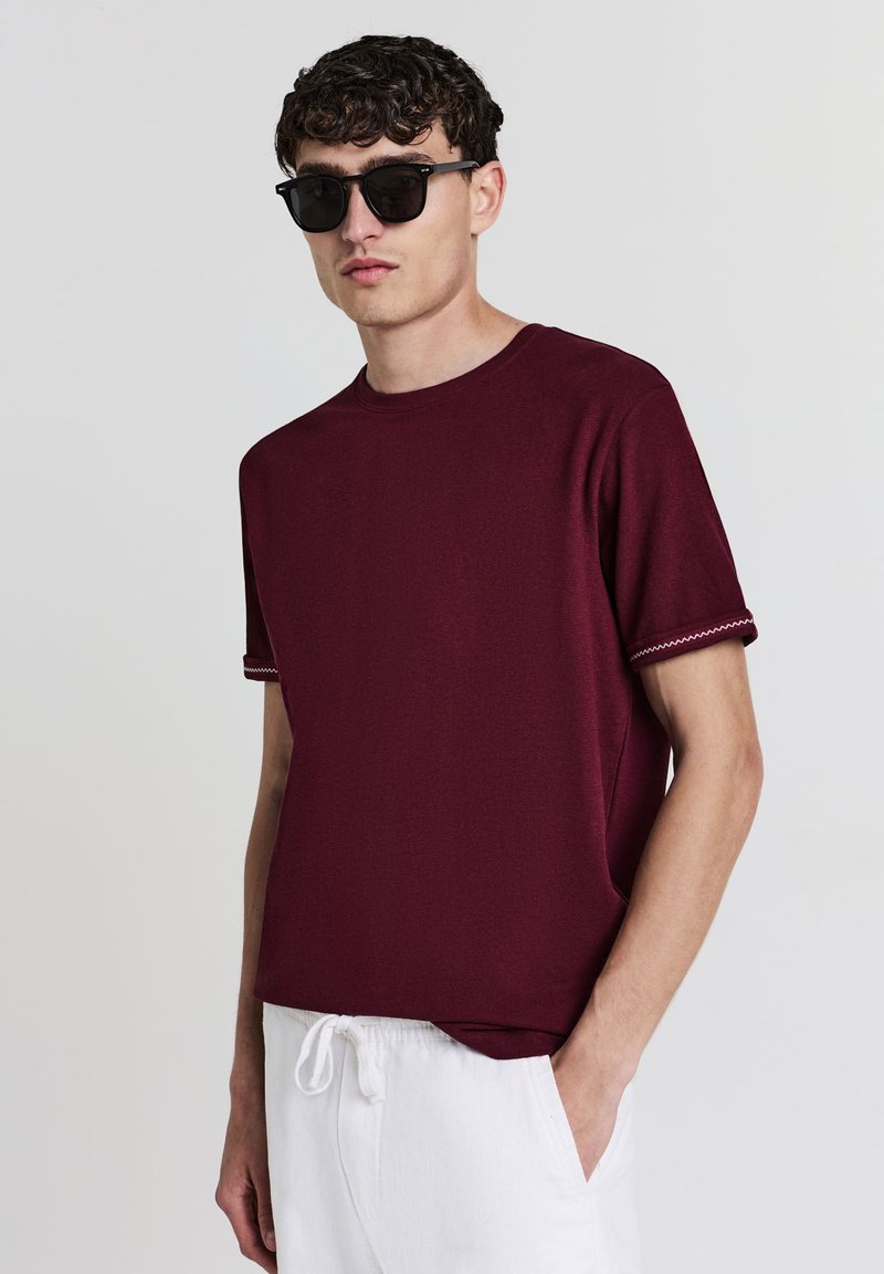 Giovane uomo con occhiali da sole scuri, una camicia a maniche corte bordeaux con cuciture bianche sulle maniche e pantaloni bianchi con cordoncino, con una mano in tasca.