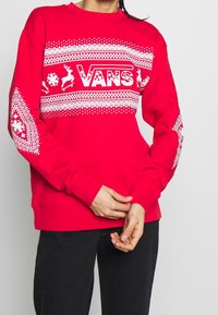 Sweatshirt rouge avec des motifs blancs sur le thème des fêtes, comprenant des rennes et des flocons de neige. Manches longues et col rond. Logo sur la poitrine.