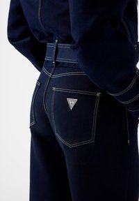 Τζιν από ναυτικό μπλε denim με λευκές ραφές. Εμφανίζει ένα τριγωνικό λογότυπο "GUESS" στην πίσω τσέπη και σχεδιασμό εφαρμοστής ζώνης μέσης.