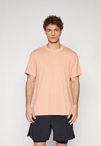 Camiseta de algodón color melocotón claro con mangas cortas, cuello redondo y un pequeño logo blanco de Nike en el lado izquierdo del pecho. Llevada con pantalones cortos negros.