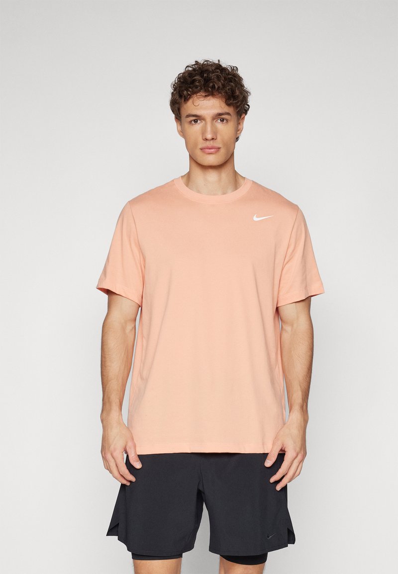 Camiseta de algodón color melocotón claro con mangas cortas, cuello redondo y un pequeño logo blanco de Nike en el lado izquierdo del pecho. Llevada con pantalones cortos negros.