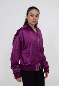 Lichtpaarse satijnen bomberjack met ribbelmanchetten en tailleband, voorzien van een voorrits en zijzakken. Gladde textuur.