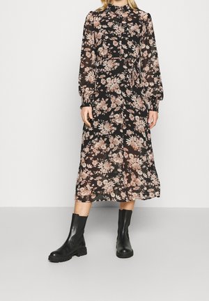 Femme portant une robe midi noire à fleurs avec manches longues et taille nouée, associée à des bottines noires, debout devant un fond uni.