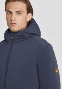 Marineblauwe zip-up jas met een hoge kraag en capuchon. Voorzien van een logopatch op de linker mouw en een gladde, gestructureerde stof.