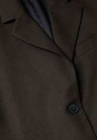 Veste de costume marron foncé avec un large revers, un seul bouton noir, et une poche poitrine sur une doublure intérieure sombre.