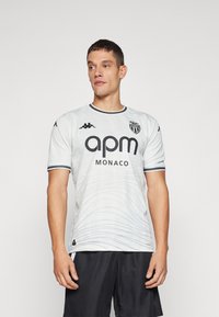 Witte voetbalshirt met korte mouwen, voorzien van een subtiele golfpatroon, zwarte accenten, groot "apm MONACO" logo, en Kappa merk op de mouwen.